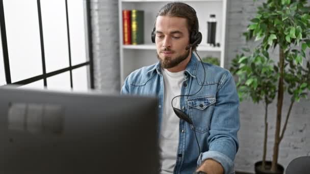 Fiatal spanyol férfi üzletember számítógépes és fülhallgató komoly arccal az irodában - Felvétel, videó