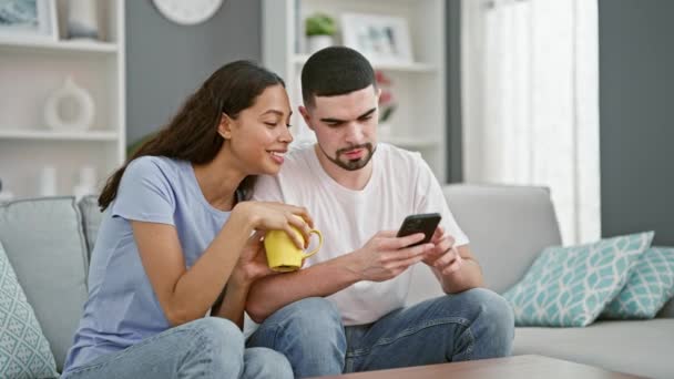Bella coppia insieme innamorati, sorridenti fiduciosi, che scrivono sullo smartphone davanti al caffè sul divano del soggiorno di casa - Filmati, video