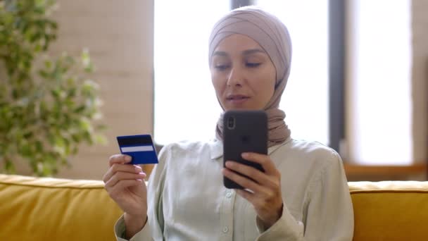 Heureuse femme du Moyen-Orient en hijab acheter des choses en ligne, en utilisant un smartphone et une carte de crédit, profiter des achats sur Internet, se reposer sur le canapé à la maison, pistage, ralenti - Séquence, vidéo