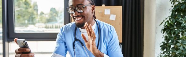 wesoły afrykański lekarz w okularach machających przed kamerą telefoniczną i uśmiechających się radośnie, sztandar - Zdjęcie, obraz