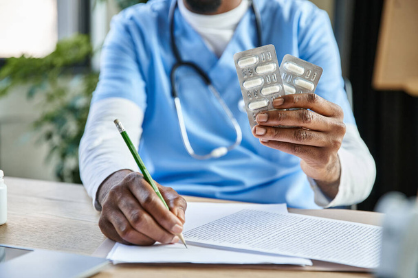 обрезанный вид африканского американского врача со стетоскопом, держащего таблетки и делающего заметки, телездравоохранение - Фото, изображение