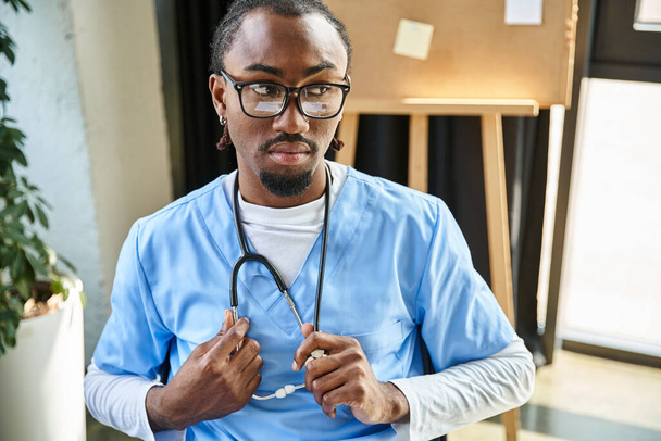 σκεπτικός όμορφος Αφροαμερικανός γιατρός με γυαλιά που κρατάει στηθοσκόπιο και κοιτάζει αλλού. - Φωτογραφία, εικόνα