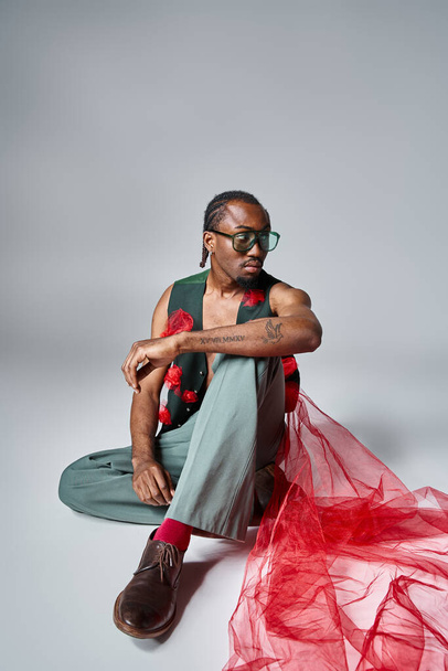 bel homme afro-américain avec du tissu de tulle rouge sur son gilet assis sur le sol, concept de mode - Photo, image