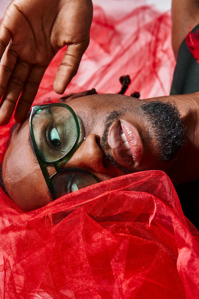 portret van knappe Afro-Amerikaanse man met rode tule stof liggend op de vloer, mode-concept - Foto, afbeelding