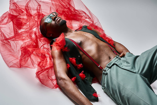 attractive young african american male model in stylish attire with red tulle fabric lying on floor - Photo, Image