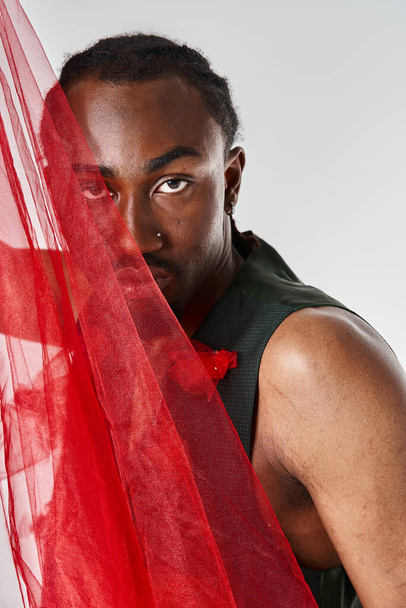 handsome young african american male model posing with vibrant red tulle fabric on gray backdrop - Photo, Image