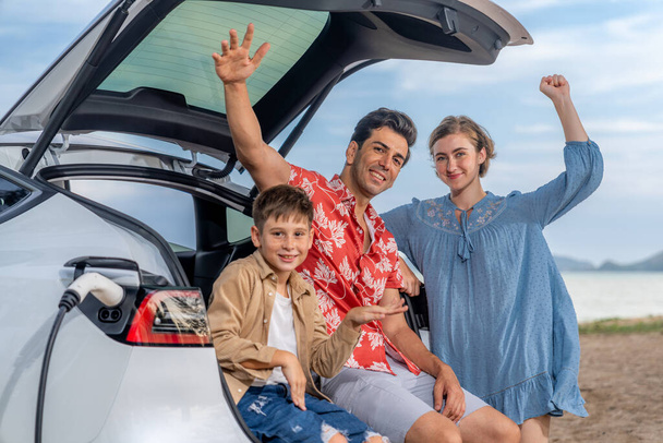 Rodzinny wyjazd wakacyjny podróżujący przez plażę z elektrycznym samochodem, piękna rodzina siedzieć na bagażniku, ładowania akumulatora samochodowego EV z zielonej i zrównoważonej energii. Podróże rodzinne i ekologiczny samochód. Wieczne - Zdjęcie, obraz