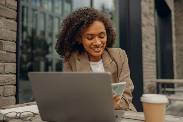 Usmívající se žena na volné noze pracující na notebooku, zatímco sedí v kavárně na terase. Koncept práce na dálku - Fotografie, Obrázek
