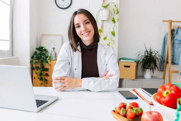 Portrait of young beautiful nutritionist woman smiling at camera at medical consultation. Professional healthcare worker concept - Photo, Image
