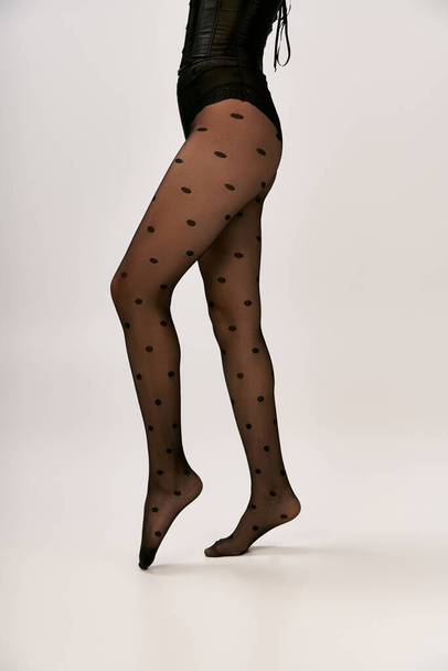 cropped photo of chic woman in black corset and polka dot tights posing on white backdrop, tip toe - Photo, Image