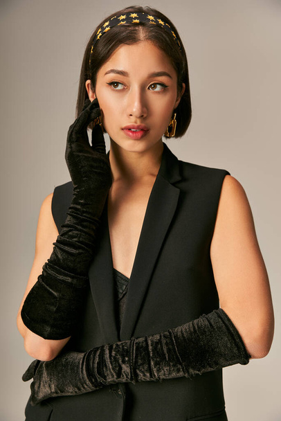 encantadora mujer asiática en elegante atuendo con guantes y diadema posando sobre fondo gris - Foto, Imagen