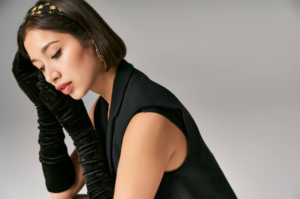 привлекательная и мечтательная азиатка в элегантной одежде с перчатками на сером фоне - Фото, изображение
