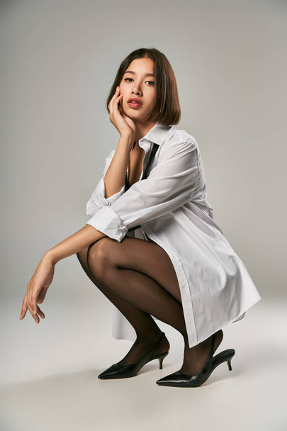teljes hossza ázsiai modell fehér ing, fekete sarkú és harisnyanadrág ül szürke háttér - Fotó, kép