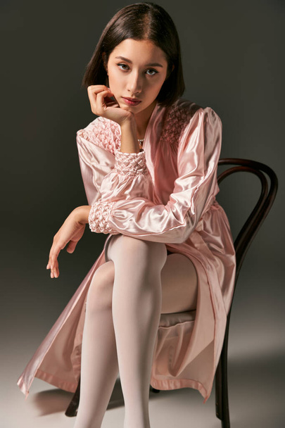 bonito jovem asiático mulher no rosa seda robe e branco collants sentado no cadeira no cinza fundo - Foto, Imagem