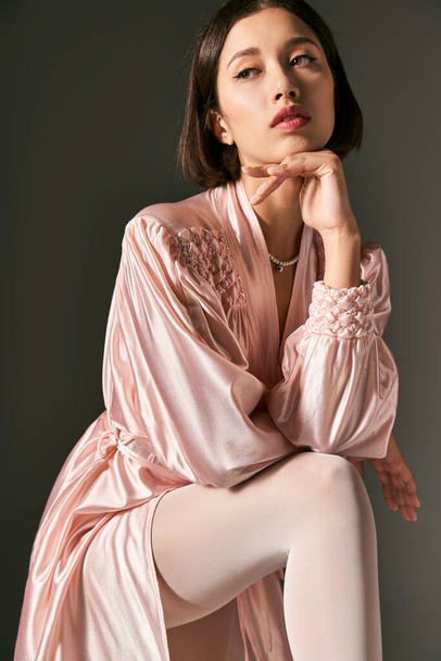 dreamy and young asian woman in pink silk robe and white pantyhose posing on grey background - Photo, Image