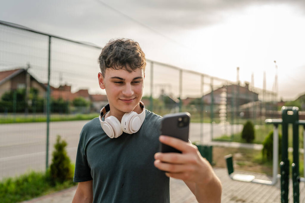 Jeden muž kavkazský mladý muž stojí na venkovním otevřeném tréninkovém parku tělocvična používat mobilní telefon smartphone se sluchátky posílat zprávy SMS nebo procházet on-line aplikace pro školení reálné osoby kopírovat prostor - Fotografie, Obrázek