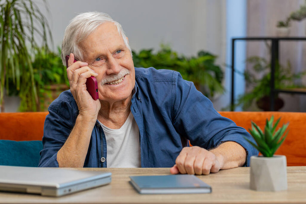 Telefonanrufe, gute Nachrichten, Klatsch. Glücklich lächelnd überraschte ein älterer Herr im angenehmen Gespräch auf dem Smartphone und genoss das Gespräch mit einem Freund zu Hause in der Wohnung. Älterer Großvater sitzt auf Couch am Tisch - Foto, Bild