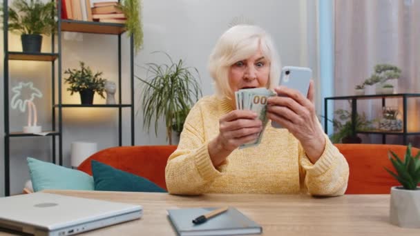 Gezinsbudget plannen. Glimlachende senior vrouw die geld telt smartphone gebruiken berekenen binnenlandse rekeningen thuis. Volwassen oude grootmoeder tevreden van inkomen en bespaart geld voor geplande vakantie geschenken - Video