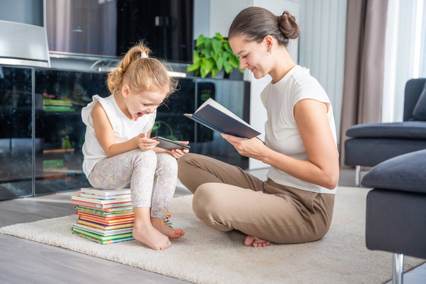 Das kleine Mädchen sitzt auf einem Stapel Kinderbücher und bedient sein Smartphone, während ihre Mutter ein Buch liest. Hochwertiges Foto - Foto, Bild