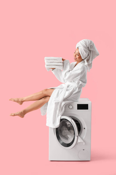 Beautiful young woman in bathrobe with stack of clean clothes sitting on washing machine against pink background - Photo, Image
