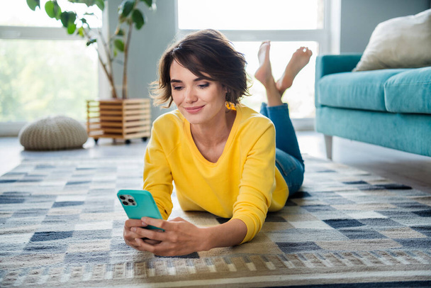 Photo of positive lady millennial lying floor using smart gadget texting in modern apartment flat. - Zdjęcie, obraz