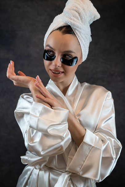 Cheerful young woman in white towel applied collagen patch for smoothing skin under eye isolated on black. Anti aging procedures - Photo, Image