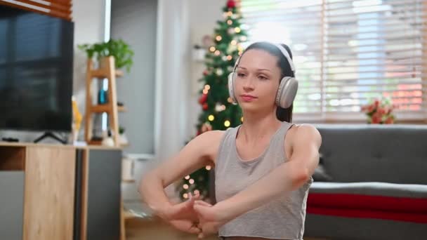 Vonzó fiatal nő fülhallgató nyújtás gyakorlatok otthon a nappaliban díszített karácsonyfa. - Felvétel, videó