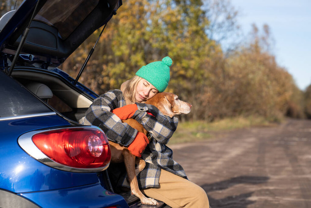 Middle aged woman in road trip with dog hugging pet sitting in car trunk during break on nature. Affectionate dog lover tenderly hugs animal with closed eyes feels harmony. Travel tourism road trip.  - 写真・画像