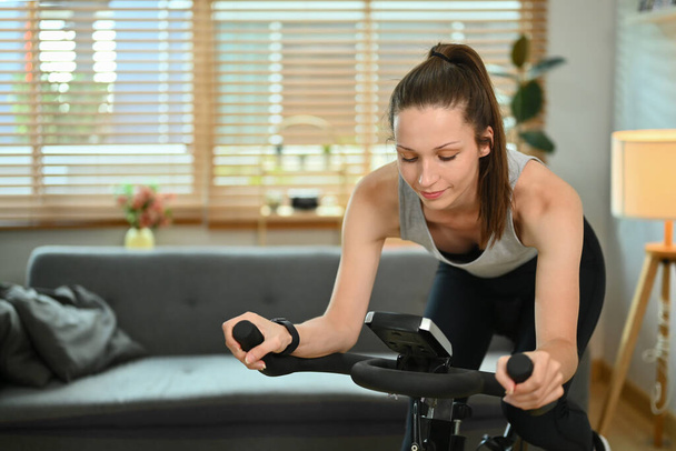 Shot van jonge sportvrouw doet cardio op stationaire fiets thuis. - Foto, afbeelding