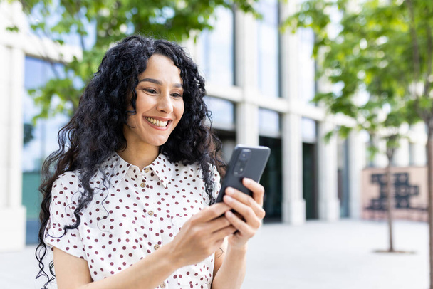 Молода красива жінка прогулюється містом з телефоном в руках, задоволено посміхається, використовує додаток для смартфона, переглядає сторінки в інтернеті, вводить текстове повідомлення, соціальні мережі. - Фото, зображення