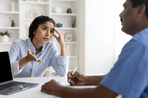 Young Indian Female Patient Sharing Health Problems With Doctor Man During Meeting In Office, Upset Sick Woman Talking To Mature Therapist, Male Physician Making Notes To Clipboard, Selective Focus - Photo, Image