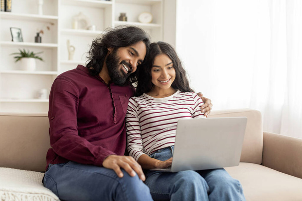 Szerető vidám indiai pár laptoppal együtt, otthonos belső tér, boldog Y-generációs férfi ölelgeti gyönyörű feleségét, és nézi a notebook képernyőn, képeket néz, másolás helyet - Fotó, kép