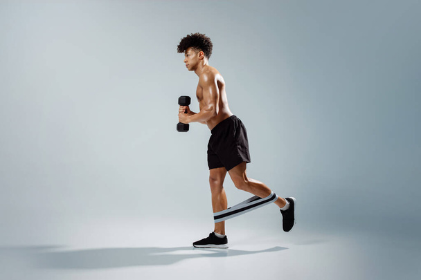 Entschlossener junger Athlet mit Kurzhanteln beim Training mit Gummischlaufe an den Beinen, hemdlos auf grauem Hintergrund stehend, muskulöse Arme und Oberkörper hervorhebend. Gym Workout und Fitness - Foto, Bild