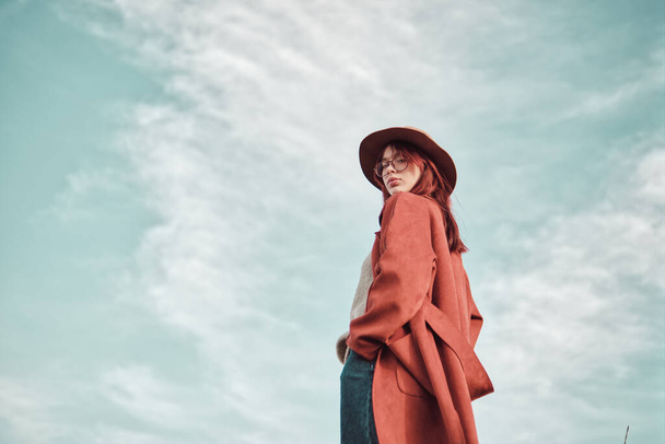 Kırmızı paltolu ve şapkalı kendine güvenen genç kız elleri ceplerinde gökyüzüne bakıyor ve omzunun üzerinden kameraya bakıyor. Düşük açı görünümü. - Fotoğraf, Görsel