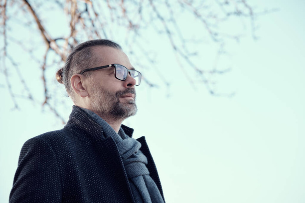 Portrait of a confident bearded middle-aged man with glasses wearing a blue coat and woollen scarf against the sky. Low angle view. - Photo, Image