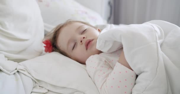 かわいい女の子の幼児は枕付きの快適なベッドで眠り,柔らかい毛布で覆われました. ベッドルームに落ち着く小さな子供のクローズアップ. ベッドタイムコンセプト。 低アレルギーベッド. - 映像、動画