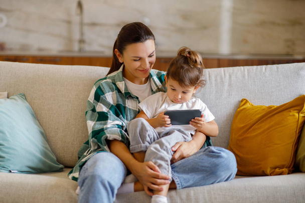 Νεαρή μητέρα και η μικρή κόρη της χρησιμοποιώντας Smartphone μαζί στο σπίτι, μαμά και χαριτωμένο μικρό παιδί βλέποντας κινούμενα σχέδια σε απευθείας σύνδεση, ενώ κάθεται στον καναπέ στο εσωτερικό του σαλονιού, αντιγραφή χώρου - Φωτογραφία, εικόνα