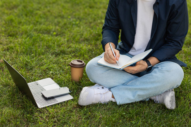 Millennial european guy enjoy lifestyle, work on laptop, phone, make notes in book, planning business, sit on grass in city, outdoor. Study, education, startup with gadget - Photo, Image