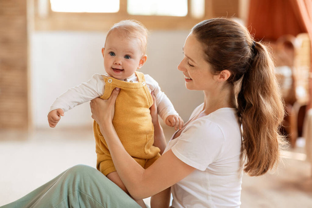 Onnellinen äiti poseeraa vauvan halaus ja kuljettaa söpö pieni lapsi käsissä istuu lattialla makuuhuoneessa kotona, Kid hymyilee kameralle. Äitiyden ilo, äitiysloma elämäntapa käsite - Valokuva, kuva