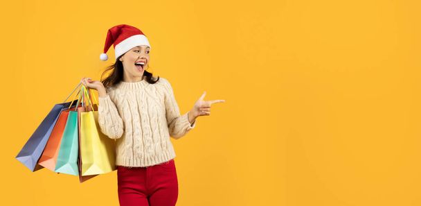 Vrouw shopaholic in feestelijke Santa hoed en met boodschappentassen in de hand balken met vreugde, wijzend vinger opzij naar de vrije ruimte, reclame winter vakantie aanbieding op gele studio achtergrond. Panorama - Foto, afbeelding