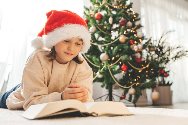 Egy Mikulás sapkás boldog lány ül egy díszített karácsonyfa mellett, és könyvet olvas. Várom az ünnepet, az új évet és a karácsonyt. - Fotó, kép