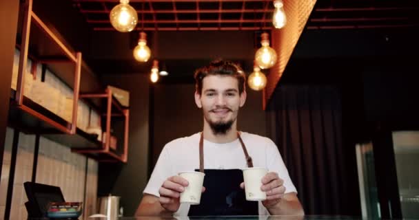 Bajo ángulo de barista barbudo joven positivo en camiseta blanca y delantal sonriendo y mirando a la cámara mientras sirve tazas de café en el mostrador en la cafetería moderna - Metraje, vídeo