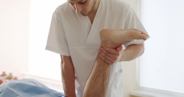 Ο Οστεοπαθητικός θεραπεύει τον ασθενή στην κλινική. Στιγμιότυπο ανδρικού θεραπευτή μασάζ που τεντώνει το πόδι του γυμνού άντρα ξαπλωμένου στο τραπέζι μασάζ κατά τη διάρκεια της συνεδρίας αποκατάστασης στην κλινική οστεοπάθειας. - Πλάνα, βίντεο