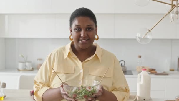 Retrato de mujer negra alegre sosteniendo tazón de cristal de ensalada de verduras, posando en la cámara y sonriendo mientras está de pie en la cocina en casa - Imágenes, Vídeo