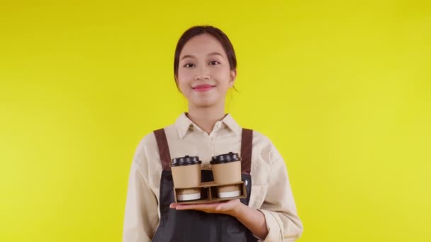 Portret młoda azjatycka barista kobieta w fartuchu trzyma filiżankę kawy i prezentuje na żółtym tle, kelnerka wesoły i pokazując i kciuki do góry, mały biznes lub startup, kelner kawiarni. - Materiał filmowy, wideo