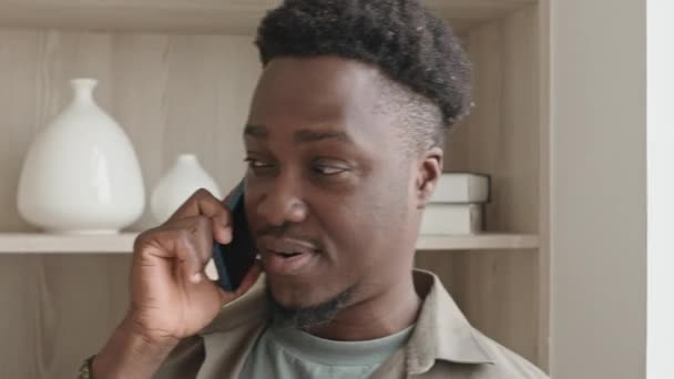 自宅の本棚の近くに立って携帯電話で話しているアフリカ系アメリカ人男性の中型のクローズアップショット - 映像、動画