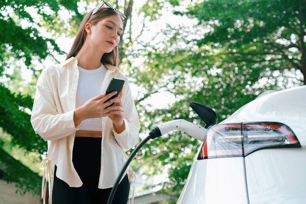 Mujer joven utiliza el teléfono inteligente para pagar la electricidad en la estación de carga de coches EV público parque verde de la ciudad. Moderno estilo de vida urbano ambiental y sostenible con vehículo EV. Expediente - Foto, imagen