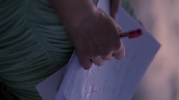 Une jeune femme en robe légère écrit une lettre sur des feuilles de papier, assise sur un rocher sur fond de mer éclairée par le coucher du soleil. Vertical, Ralenti, Gros plan. - Séquence, vidéo