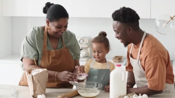 Allegro afroamericano madre sbattere gli ingredienti alimentari in ciotola mentre suo marito gioca con divertente figlia bambino durante il tempo di cottura in cucina - Filmati, video