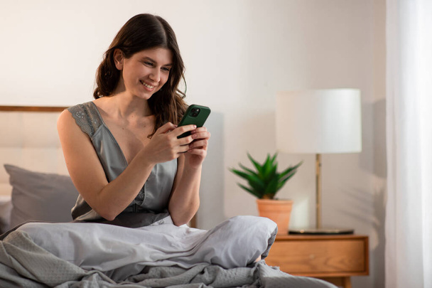 Menina sorridente está sentado na cama com smartphone pela manhã. Jovem feliz digitando uma mensagem telefônica depois de acordar. Foto de alta qualidade - Foto, Imagem
